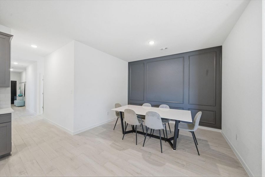 This photo showcases a modern dining area with a sleek white table and six chairs. It features a dark accent wall, light wood flooring, and an open hallway leading to other spaces.