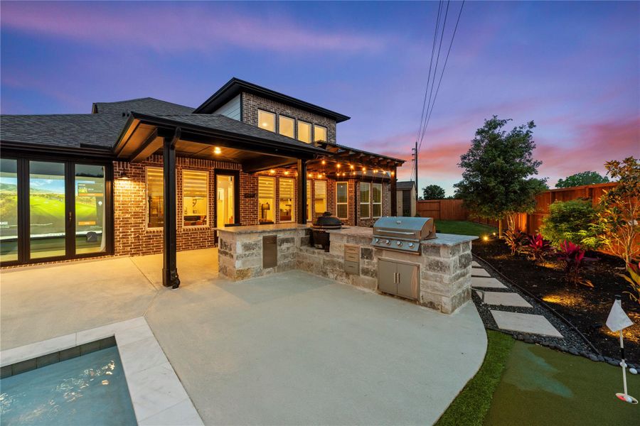 A view of the Texas-sized covered patio complete with ambient lighting and an outdoor kitchen. The area features a small putting green, landscaped garden, and overlooks a serene pool, perfect for outdoor entertainment and relaxation.