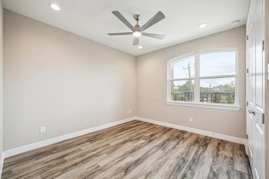 This is a bright, unfurnished room featuring modern wood-look flooring, a contemporary ceiling fan, and a large window allowing for plenty of natural light. The neutral color palette offers a versatile backdrop for personalization.