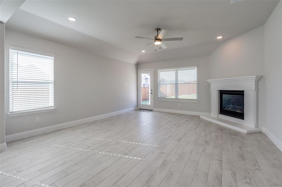 Spacious living area featuring wood-finish flooring, a corner fireplace with a white mantel, multiple windows with blinds, a ceiling fan, and recessed lighting