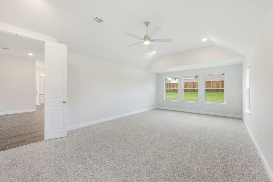 Spacious bedroom with vaulted ceiling, ceiling fan, carpet flooring, and three windows overlooking backyard