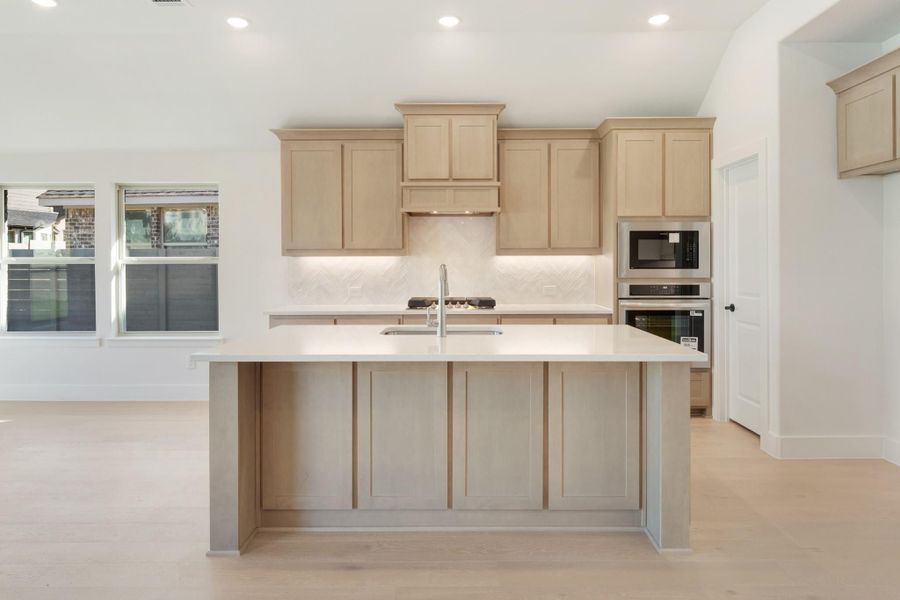 Kitchen with large center island, light wood cabinets, herringbone backsplash, and stainless steel double ovens