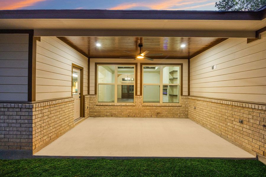 The spacious covered patio with brick and siding walls, recessed lighting, and a ceiling fan, perfect for outdoor relaxation or entertaining. Large windows offer a view into the home, and the surrounding greenery adds a touch of nature.