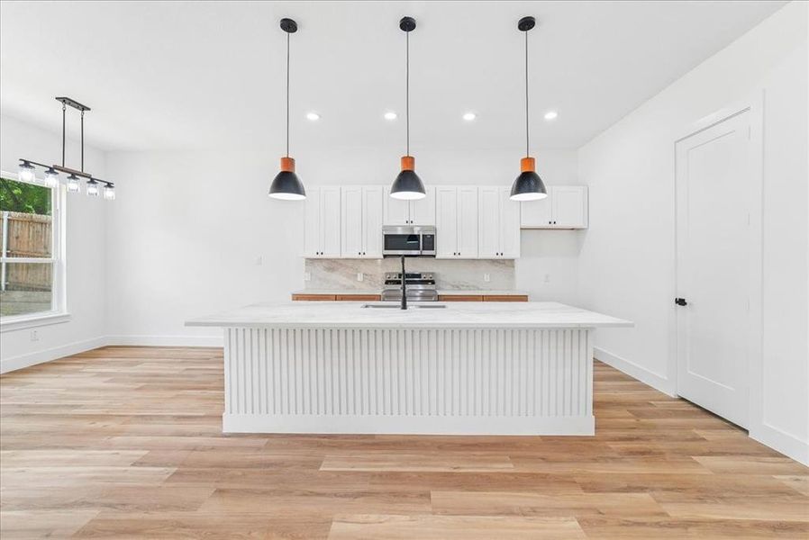 Spacious kitchen featuring a large island with a fluted panel base and a light-toned countertop, white cabinetry with dark hardware, a stainless steel microwave, and wood-finish flooring Spacious kitchen featuring a large island with a fluted panel base and a light-toned countertop, white cabinetry with dark hardware, a stainless steel microwave, and wood-finish flooring