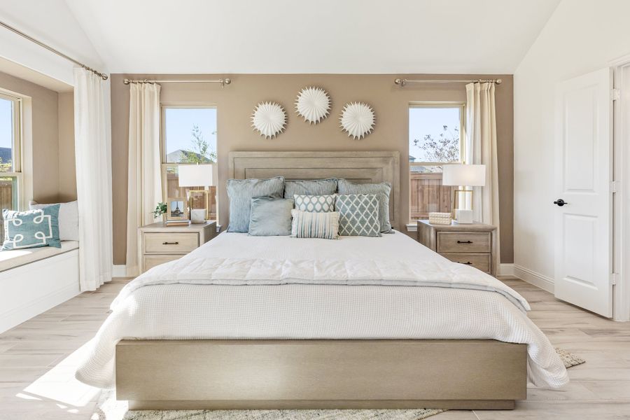Master bedroom with king bed, light wood floors, window seat, and two windows with white curtains