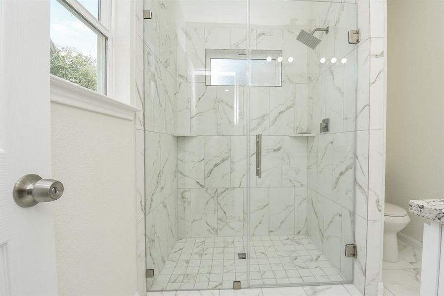 Modern bathroom with marble-tiled walk-in shower, glass door, window, and toilet visible. White and clean design with overhead and vanity lighting.