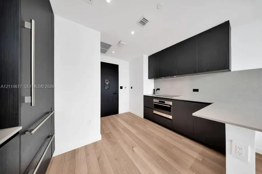 Kitchen featuring flat-panel dark cabinetry, integrated refrigerator, neutral solid-surface countertops, recessed lighting, and wood-finish flooring Kitchen featuring flat-panel dark cabinetry, integrated refrigerator, neutral solid-surface countertops, recessed lighting, and wood-finish flooring