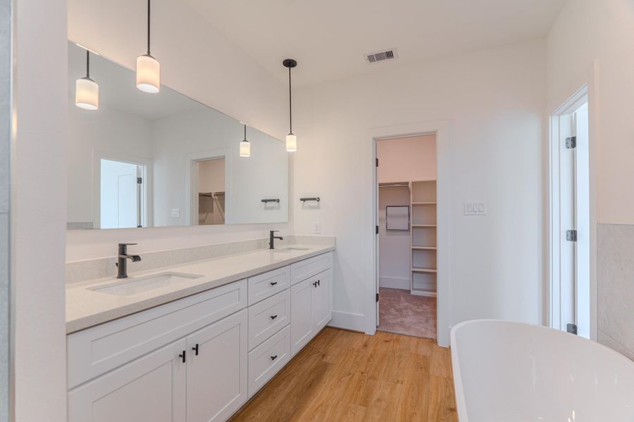 This is a modern bathroom with dual sinks, white cabinetry, and pendant lighting. It features a large mirror, wood flooring, and access to a walk-in closet.Photo Disclaimer: Photos are representative of the community and may depict a similar floor plan, model home, or previously completed residence. This is a modern bathroom with dual sinks, white cabinetry, and pendant lighting. It features a large mirror, wood flooring, and access to a walk-in closet.Photo Disclaimer: Photos are representative of the community and may depict a similar floor plan, model home, or previously completed residence.