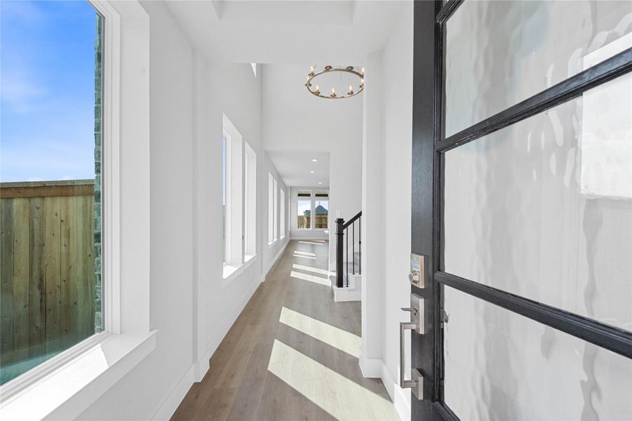 Step inside to a bright interior entryway. The space is accentuated by high, vaulted ceilings and a contemporary, round multi-light chandelier. The hallway extends toward large windows at the rear, flooding the area with natural light.