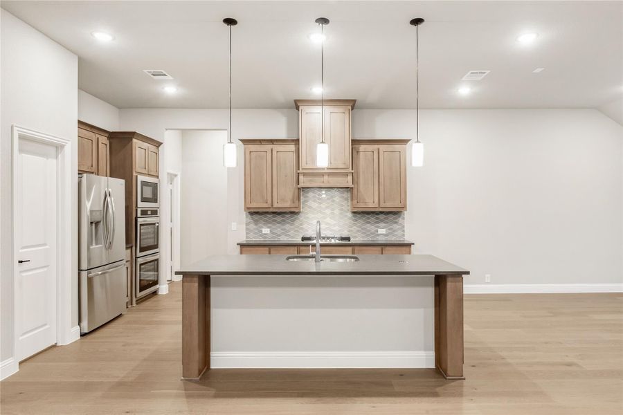 Kitchen with light wood cabinets, center island with sink, pendant lights, and stainless steel appliances on light hardwood floors