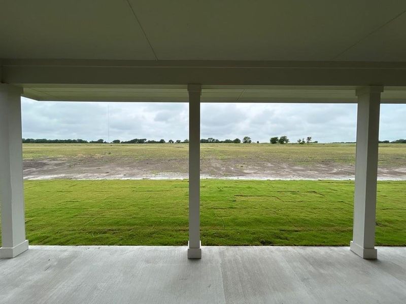 Covered patio with a concrete surface, supported by white columns, overlooking a partially sodded lawn and expansive open fields