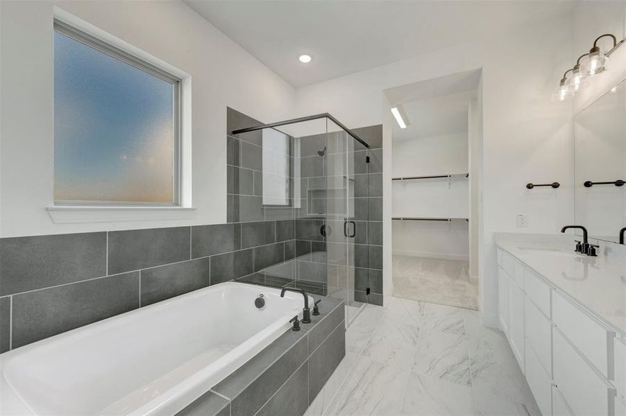 Spacious bathroom featuring a built-in tub with dark tile surround, a glass-enclosed shower, dual vanity with white cabinetry and light-colored countertops, and marble-look floor tile Spacious bathroom featuring a built-in tub with dark tile surround, a glass-enclosed shower, dual vanity with white cabinetry and light-colored countertops, and marble-look floor tile