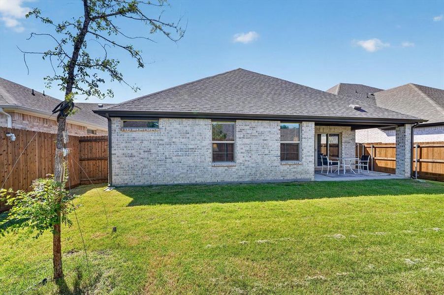 Expansive green lawn with a covered patio, featuring a brick exterior with multiple windows, a dark shingle roof, and wood fencing Expansive green lawn with a covered patio, featuring a brick exterior with multiple windows, a dark shingle roof, and wood fencing