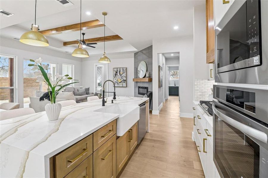 Open-concept kitchen featuring a large island with a waterfall countertop and farmhouse sink, gold-toned pendant lighting, integrated stainless steel appliances, and light wood-finish flooring
