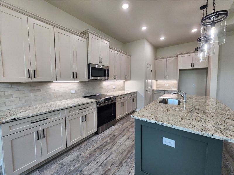 Kitchen featuring a central island with a built-in sink, stone countertops, light cabinetry with dark hardware, stainless steel appliances, and wood-finish flooring