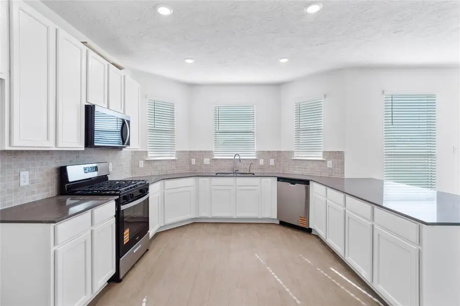This spacious kitchen boasts high ceilings, White Woodmont Belmont cabinets, Silestone Grey Expo countertops, stainless steel Whirlpool appliances, a modern Daltile backsplash, recessed lighting, windows with blinds, an added breakfast bar to kitchen countertop, and a pantry—all overlooking the expansive Great Room.