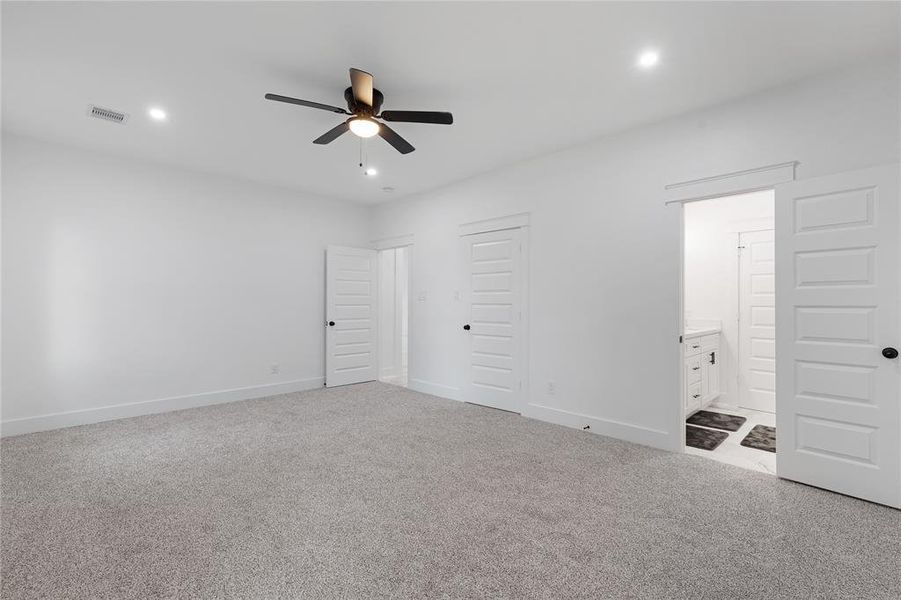 This is a spacious, well-lit room featuring neutral walls, carpeted flooring, a modern ceiling fan, recessed lighting, and two doors leading to a closet and an en-suite bathroom.