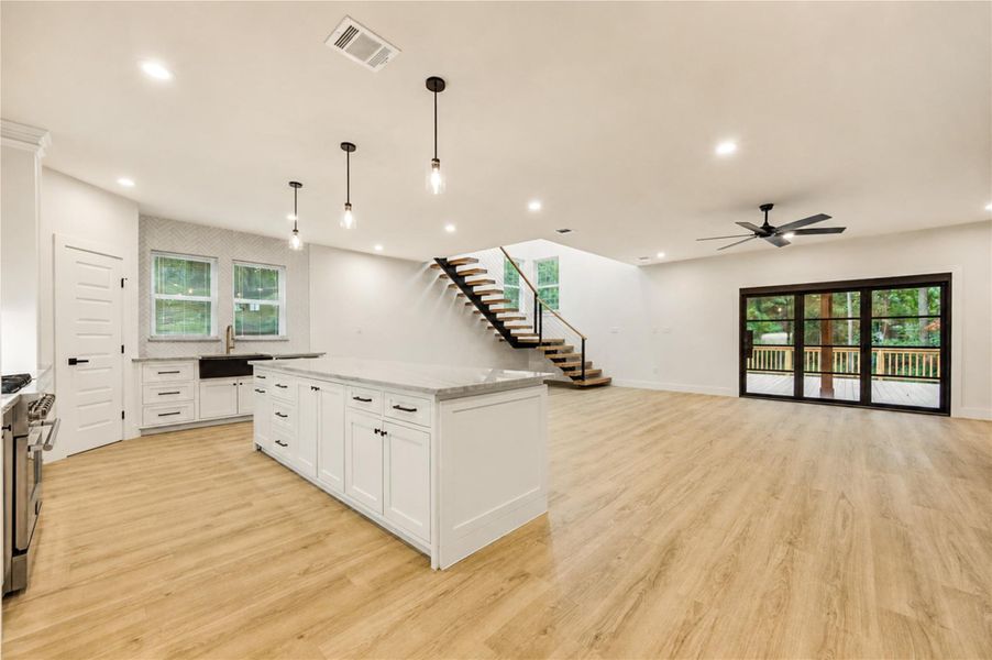 Discover a spacious open-concept living area featuring a sleek modern kitchen with a large island, elegant pendant lighting, and stylish custom cabinetry. The room seamlessly flows into a bright living space with expansive glass doors leading to a serene outdoor deck. Contemporary finishes and a striking staircase add to the home's inviting ambiance.