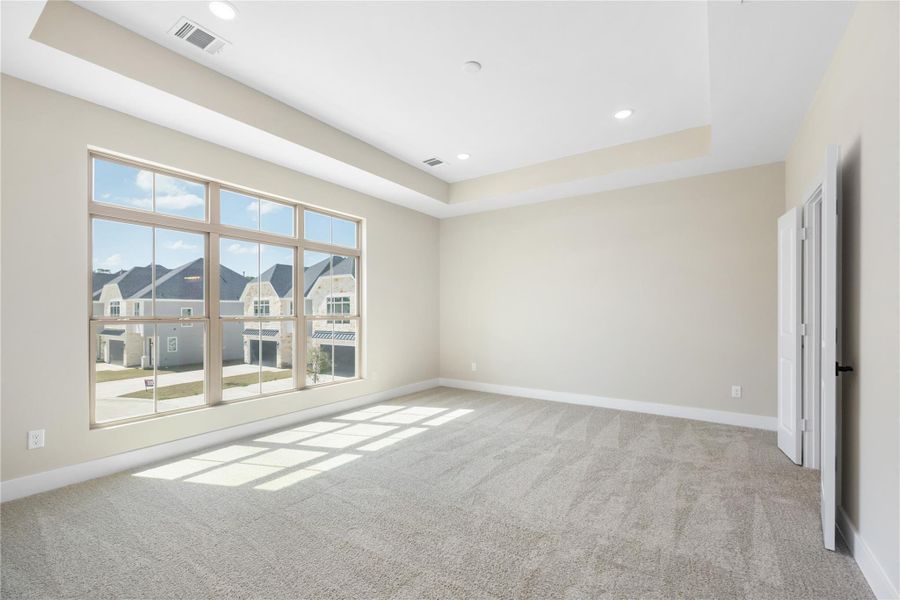 As you enter the spacious primary bedroom, you're greeted by soft carpeting and neutral walls that create a cozy and inviting atmosphere. Natural light floods the room through multiple windows, and a view into the adjacent space with large windows adds to the open feel. The elegant tray ceiling enhances the room's character, making it the perfect retreat, whether for rest, relaxation, or personalization.