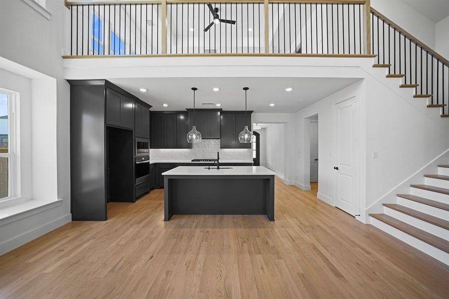 Kitchen featuring dark cabinets, pendant lighting, an island with sink, light wood-style flooring, and a high ceiling Kitchen featuring dark cabinets, pendant lighting, an island with sink, light wood-style flooring, and a high ceiling
