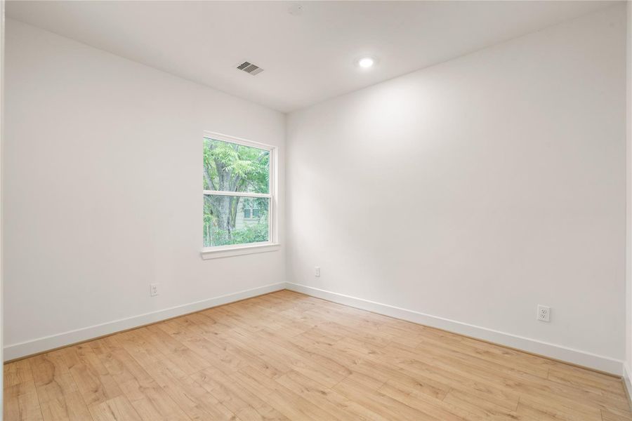 Quiet corner bedroom — warm oak-look flooring, window overlooking mature greenery, and clean neutral paint finishes.