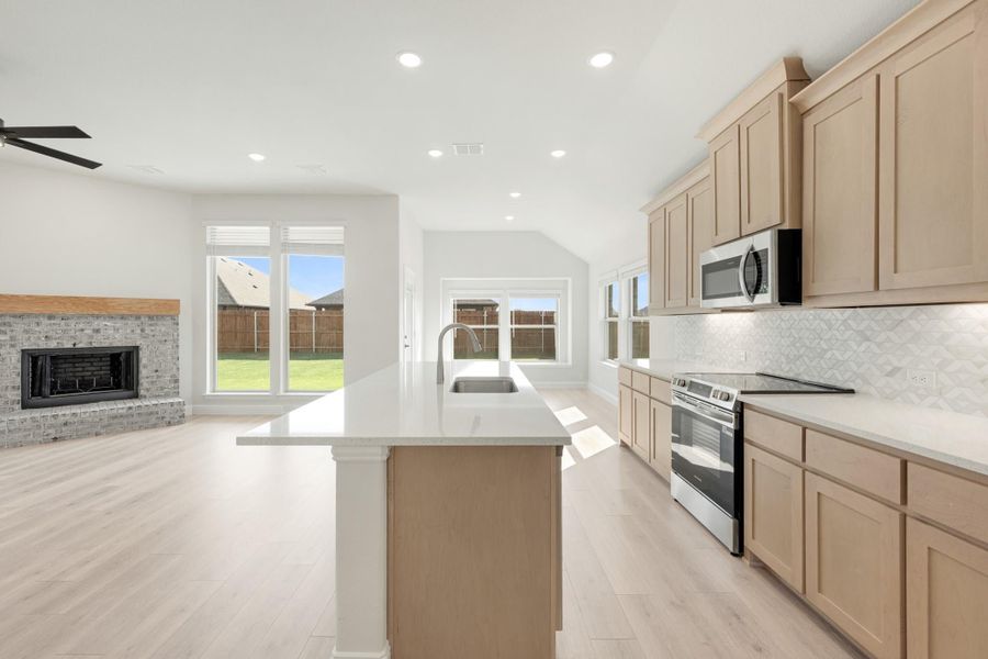 Open kitchen with light wood cabinets, center island with sink, and stainless appliances, open to living room with brick fireplace.