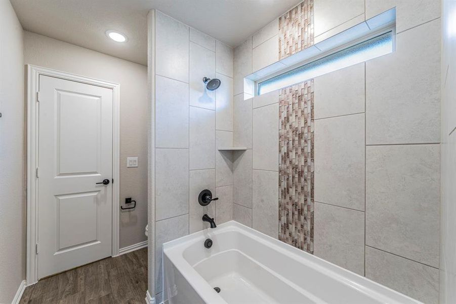 Bathtub and shower combination featuring large format tile surround, decorative mosaic tile accents, matte black fixtures, and a recessed window