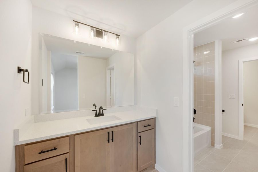 Bathroom with wood vanity, white countertop, large mirror, and tiled soaking tub in background