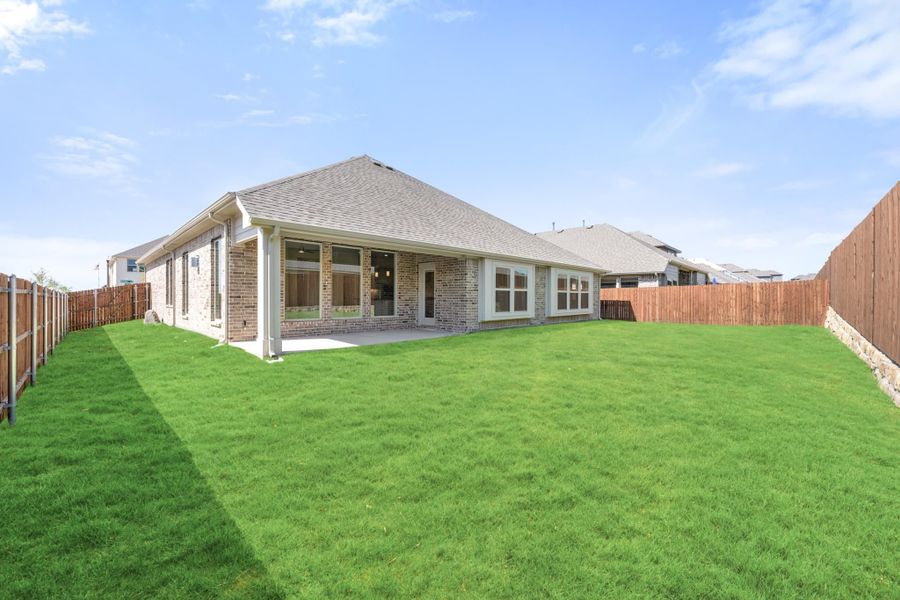 Rear view of brick home with covered patio, large grassy backyard, and wood privacy fence