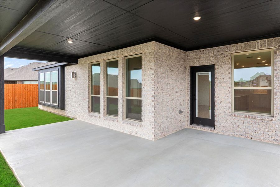 Covered back patio with concrete slab, light brick exterior, black-trimmed windows, and small fenced backyard with green grass. Covered back patio with concrete slab, light brick exterior, black-trimmed windows, and small fenced backyard with green grass.