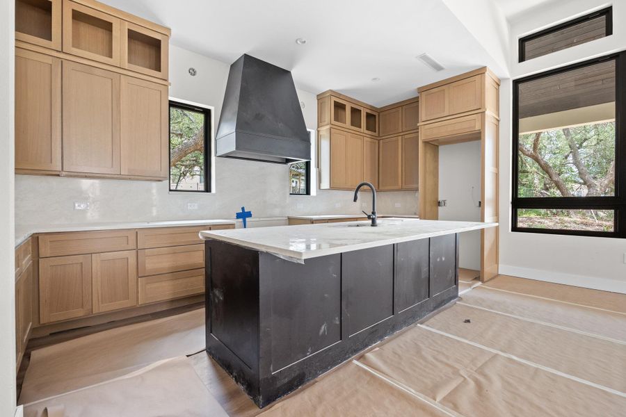 Modern kitchen featuring a large central island with a contrasting dark base, light-toned cabinetry, a sleek black range hood, a white tile backsplash, and oversized windows