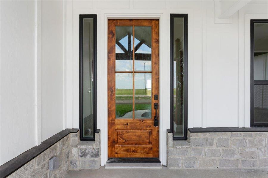 A welcoming wooden front door with clear glass panels invites entry, flanked by sleek vertical windows and a stone and white siding exterior.