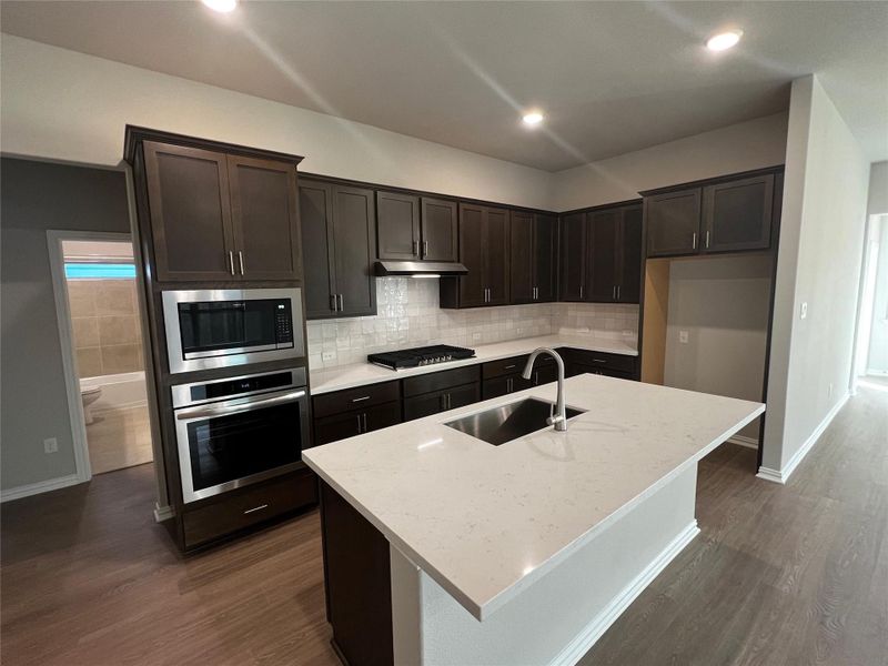 Modern kitchen featuring dark wood cabinetry, stainless steel appliances, a white subway tile backsplash, and a large island with a sink and light-colored countertop