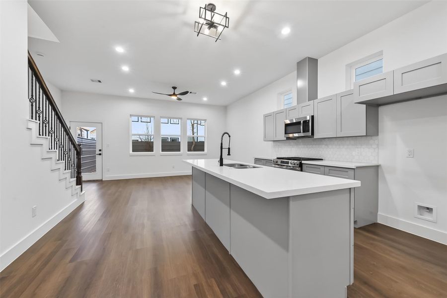 Modern open-concept kitchen and living area with sleek gray cabinetry, a large island, and hardwood floors. Features recessed lighting, stainless steel appliances, and a ceiling fan. Adjacent to a staircase and has ample natural light from large windows.