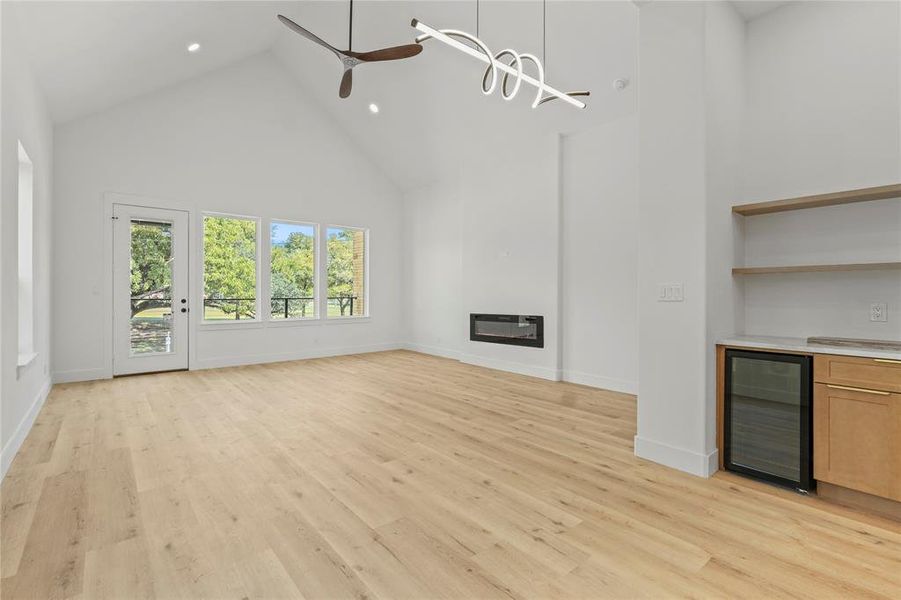 Unfurnished living room featuring high vaulted ceiling, wine cooler, a glass covered fireplace, light wood finished floors, and a ceiling fan