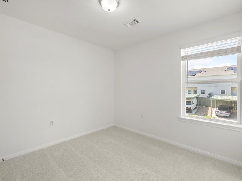A well-lit bedroom with soft carpeting and a window offering views of the neighborhood.
