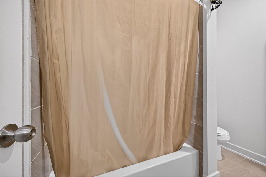 This image shows a bathroom with a bathtub and a beige shower curtain. The floor is tiled, and part of a toilet is visible, indicating a compact yet functional space. This image shows a bathroom with a bathtub and a beige shower curtain. The floor is tiled, and part of a toilet is visible, indicating a compact yet functional space.