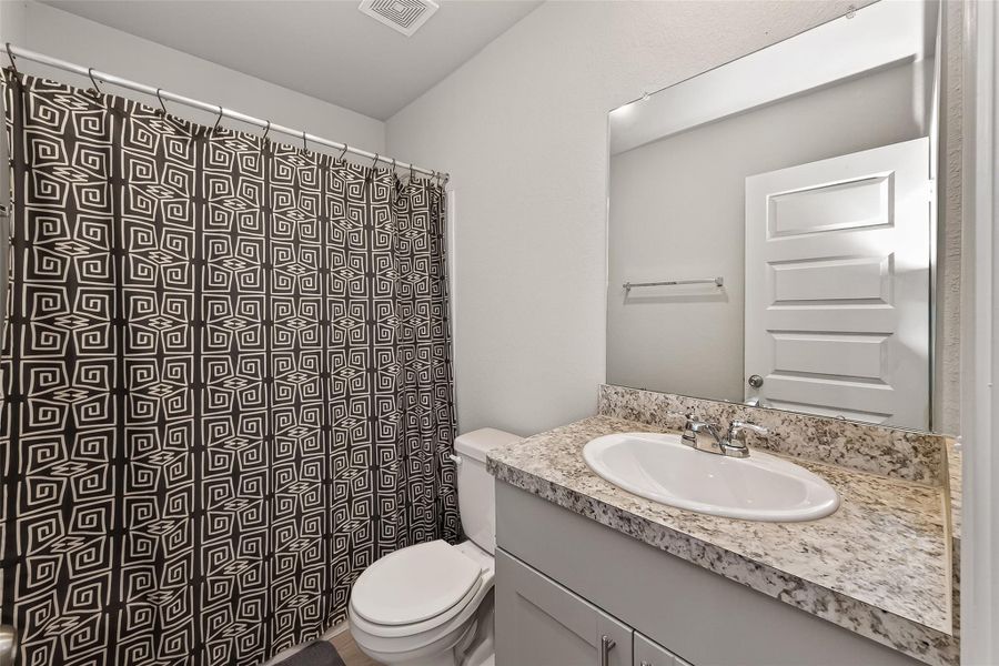 This bathroom features a modern vanity with a granite countertop and an integrated sink, complemented by a large mirror. It has a shower-tub combo with a stylish geometric-patterned curtain, offering a clean and contemporary look.