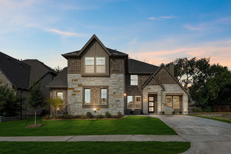 Two-story home with stone and brick exterior, dark roof, front lawn, and concrete driveway at dusk