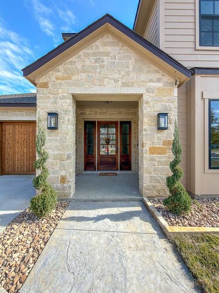 Stone facade entry featuring a solid wood front door with glass inserts, two sconce lights, a stamped concrete walkway, and landscaped rock beds with spiral topiaries