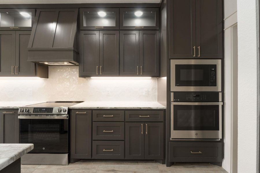 Kitchen featuring appliances with stainless steel finishes, custom exhaust hood, glass insert cabinets, decorative backsplash, and light stone countertops