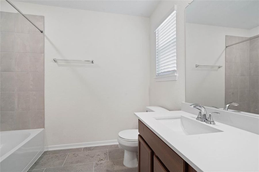 Secondary bath features tile flooring, bath/shower combo with tile surround, dark wood cabinets, beautiful light countertops, mirror, dark, sleek fixtures and modern finishes.