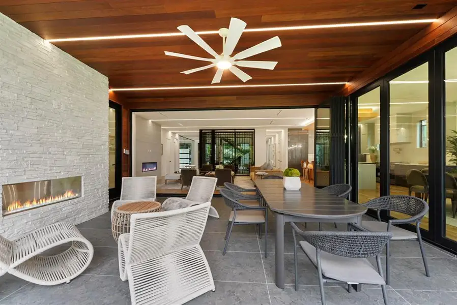Covered outdoor living area with a linear fireplace set into a stacked stone wall, wood-paneled ceiling with recessed lighting and ceiling fan, and large-format tile flooring