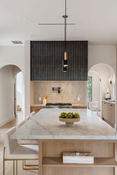 A statement tiled vent hood with an integrated pot filler anchors the kitchen, symmetrically balanced by crafted arched doorways. The island features seamlessly integrated open display shelving. A statement tiled vent hood with an integrated pot filler anchors the kitchen, symmetrically balanced by crafted arched doorways. The island features seamlessly integrated open display shelving.