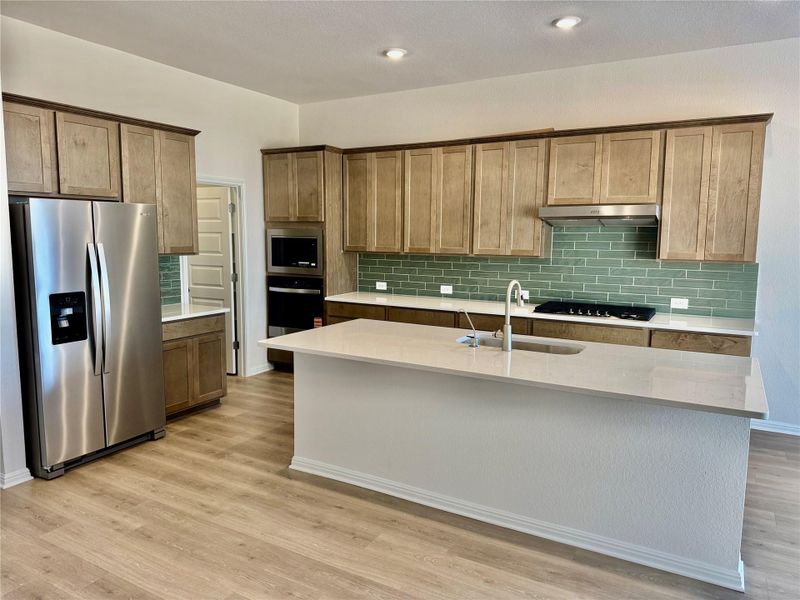 Kitchen featuring stainless steel appliances, light wood-type flooring, tasteful backsplash, light stone counters, and an island with sink