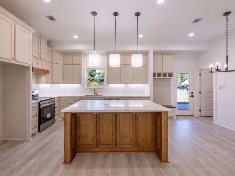 This photo showcases a modern kitchen with light cabinetry, a spacious island with a wooden base, and sleek countertops. The room is well-lit with pendant and recessed lighting, featuring stainless steel appliances and a view of the outdoors through a rear door. The open layout and light flooring create an inviting, airy atmosphere.
