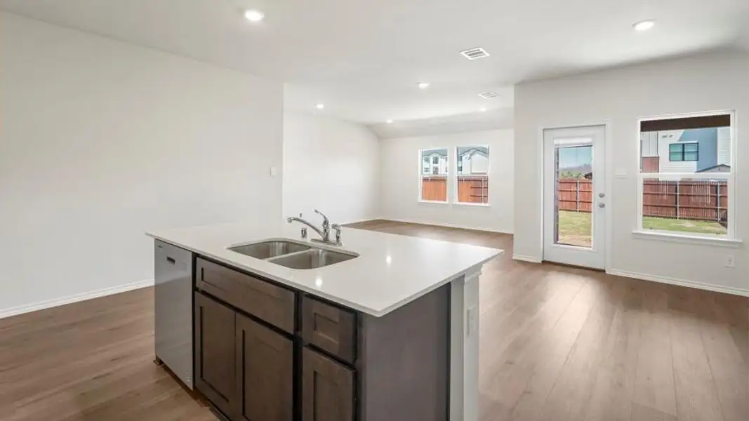 Kitchen island featuring a double-basin stainless steel sink with a gooseneck faucet, light-toned countertop, dark wood-finish cabinetry, and an integrated dishwasher