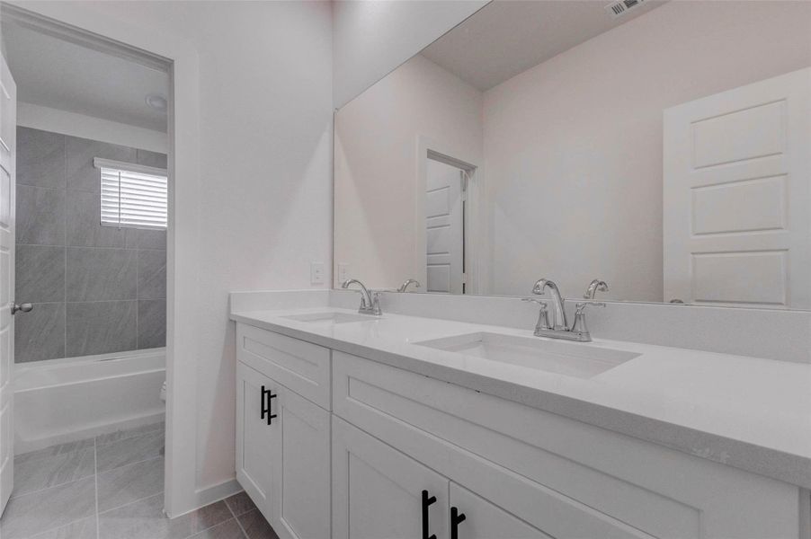 Secondary bathroom with a long double-sink vanity, large mirror, white cabinetry, black hardware, gray tile flooring, and a separate tub/shower area. The dual sinks add everyday convenience, while the neutral finishes and clean design create a fresh, functional space for bedrooms, guests, or shared use. Secondary bathroom with a long double-sink vanity, large mirror, white cabinetry, black hardware, gray tile flooring, and a separate tub/shower area. The dual sinks add everyday convenience, while the neutral finishes and clean design create a fresh, functional space for bedrooms, guests, or shared use.