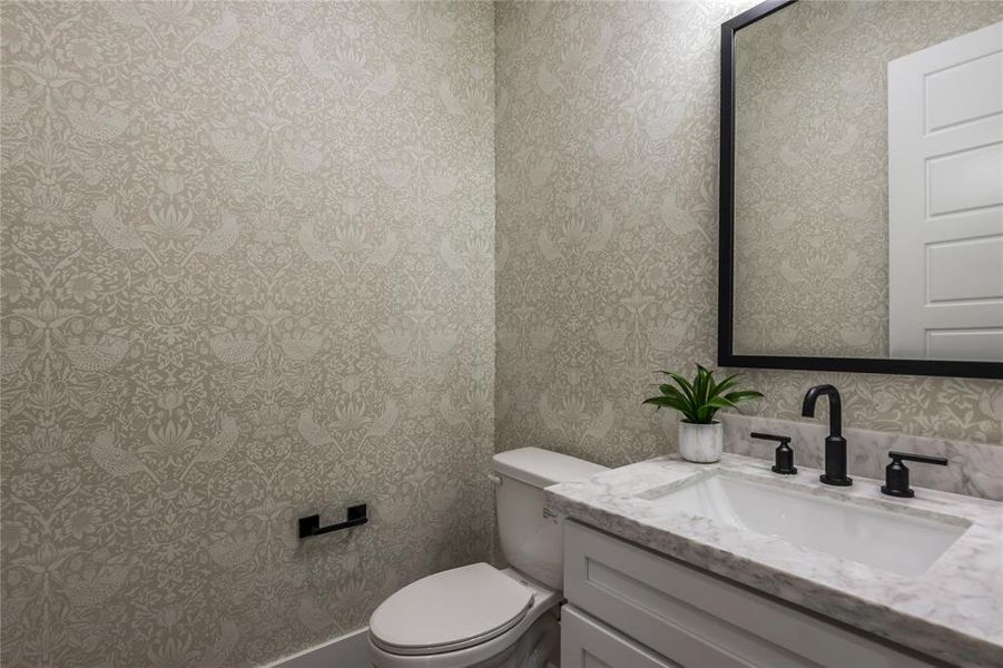 This half bathroom features elegant patterned wallpaper, a sleek vanity with a marble countertop, modern black fixtures, and a large, framed mirror, creating a stylish space. This half bathroom features elegant patterned wallpaper, a sleek vanity with a marble countertop, modern black fixtures, and a large, framed mirror, creating a stylish space.