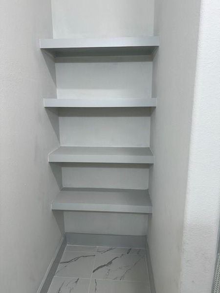 The photo shows a modern, built-in corner shelving unit with five levels, providing practical storage space in a clean, white finish that complements the neutral wall color and marble-like tile flooring.